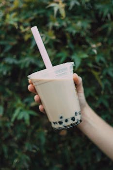 Close-up of a hand holding iced milk tea with boba pearls and straw against a leafy background.